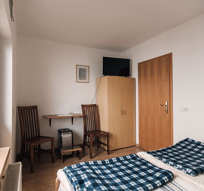 The room features two beds dressed in plaid blankets, accompanied by wooden nightstands. A simple wooden table with two chairs offers a cozy workspace. A cabinet and television are positioned against the wall, with a door providing access to the hallway.