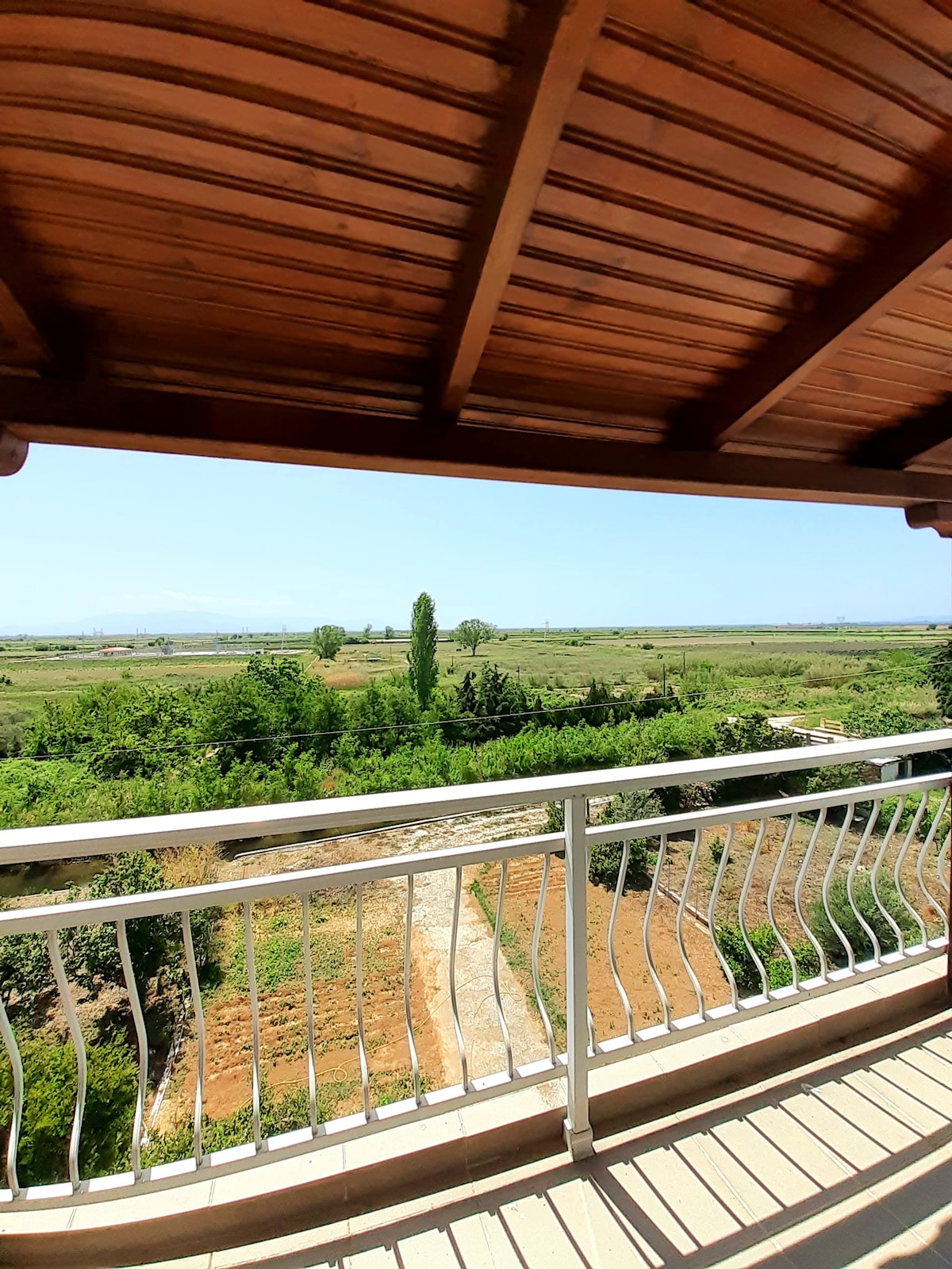 A spacious balcony showcases a stunning view of lush greenery and distant hills under a clear blue sky. The protective wooden overhang creates a shaded area for relaxation, while the metal railing provides safety. Nature's tranquility is clearly visible in the surrounding landscape.