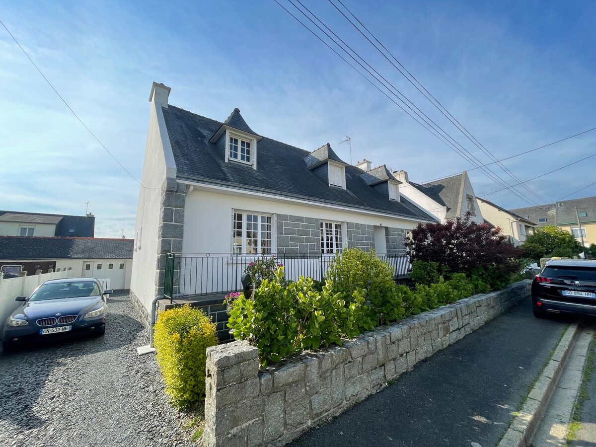 The exterior of a charming house is presented, featuring a white facade with stone accents. A sloped roof complements a front balcony surrounded by greenery. The driveway accommodates vehicles, while a well-maintained stone wall lines the pathway leading to the entrance.