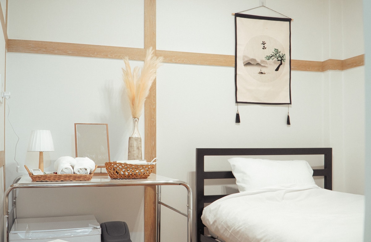 A minimally designed room features a spacious bed with crisp white bedding. A decorative wall hanging adds a subtle touch, complemented by a small table lamp and a framed mirror. A basket of towels is neatly arranged on a metallic cart, enhancing the overall simplicity.