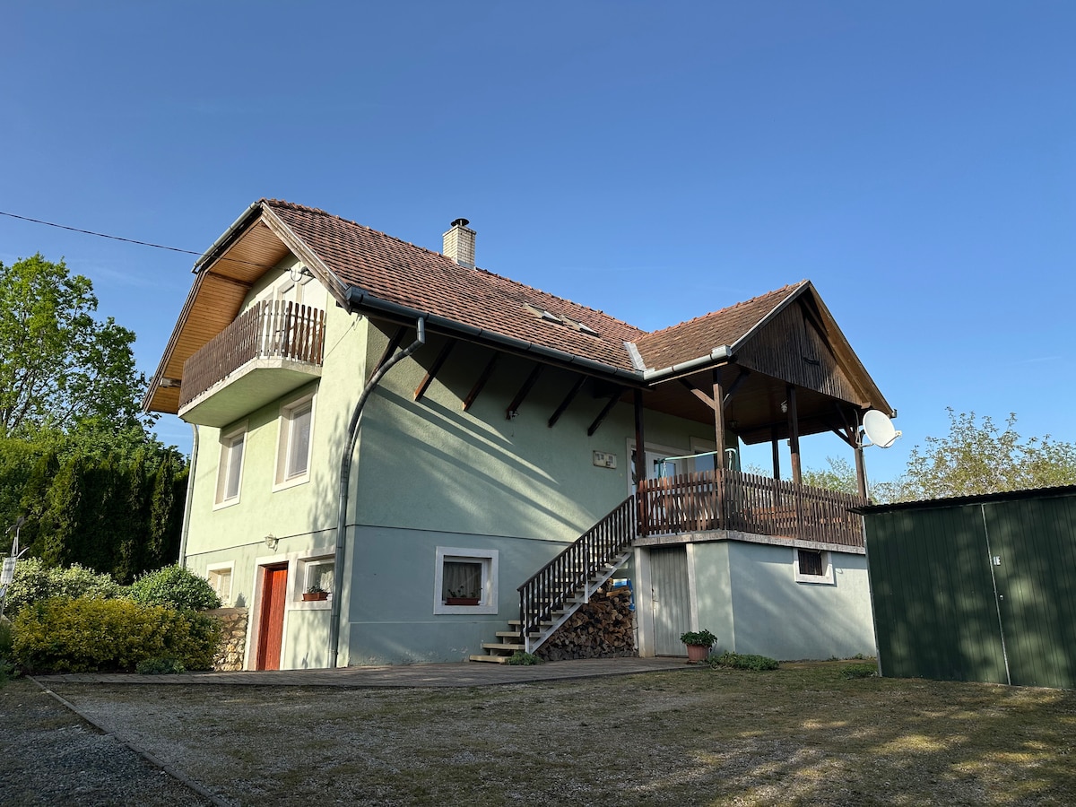 A two-story guesthouse is shown, featuring a light green exterior and a sloped roof. A spacious balcony is visible on the upper level, with wooden railings lining the stairs leading to the entrance. A well-kept garden area surrounds the house.