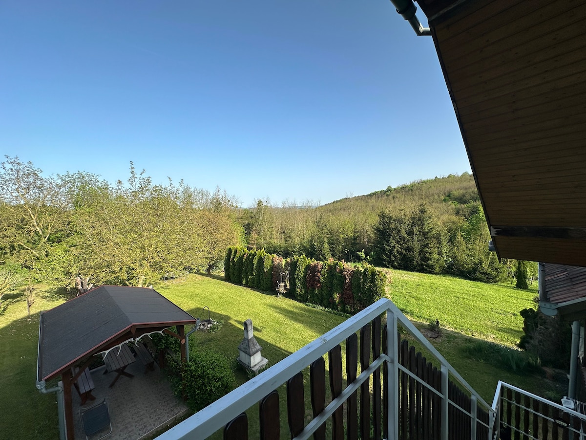 A panoramic view of a spacious garden features lush greenery and well-maintained hedges. A covered patio sits in the foreground, accompanied by a stone statue, inviting outdoor relaxation amidst the peaceful surroundings of nature.