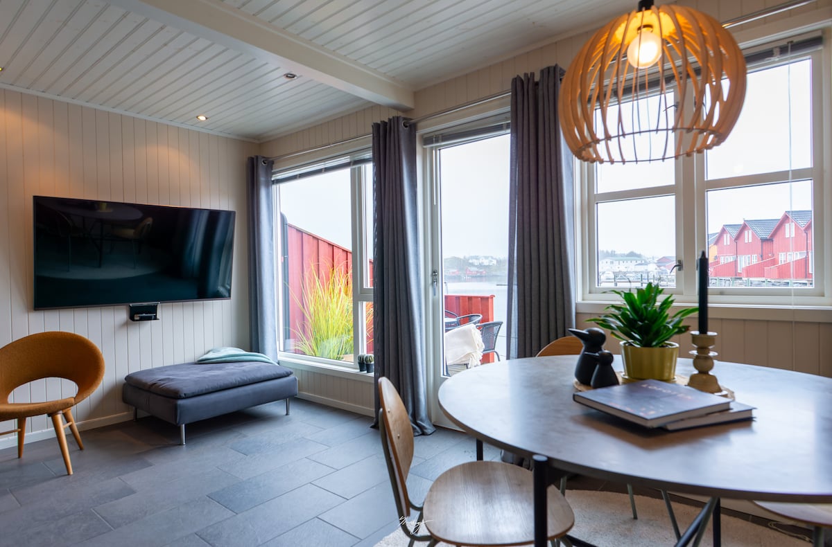 A cozy living area features a round dining table surrounded by wooden chairs. A modern gray sofa lies against the wall, and a wall-mounted TV is visible. Natural light streams in through large windows adorned with curtains, providing views of the surroundings.