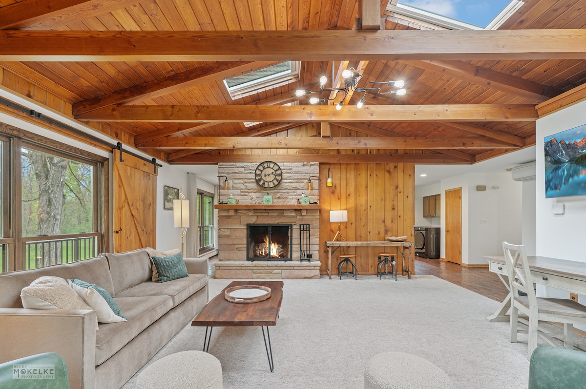 A spacious living area features a wooden beam ceiling and a stone fireplace as the focal point. Natural light floods through multiple windows, and a comfortable sofa is positioned around a coffee table. Adjacent, a dining area is visible, showcasing a rustic wooden finish.