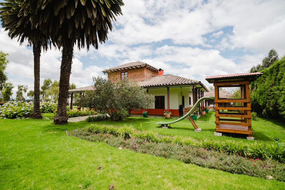 A spacious garden is surrounded by lush greenery and palm trees, featuring a play structure for children. The house displays a rustic facade with terracotta tiles and wide eaves, complemented by vibrant flowerbeds. A gentle slope leads to an area for outdoor activities.