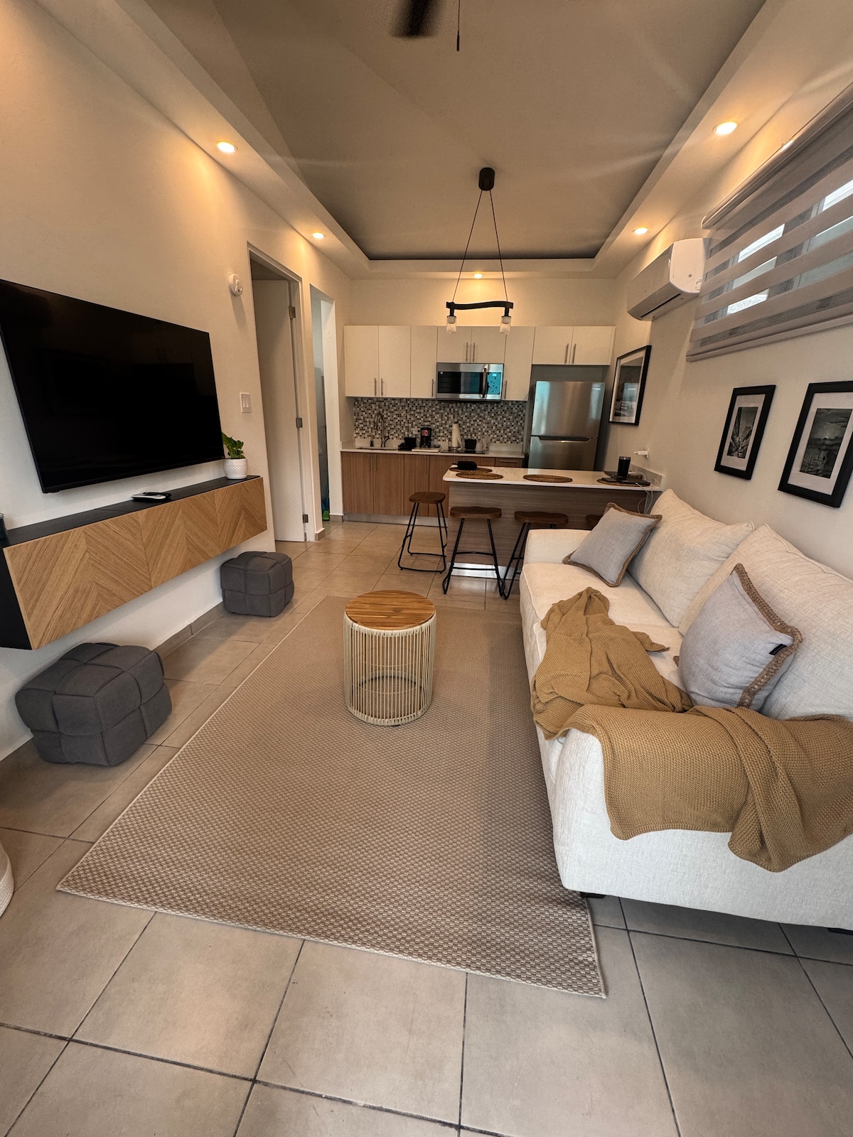 A modern living space features a light-colored sofa with a textured throw, positioned opposite a wall-mounted television. A dining area is visible in the background, showcasing a wooden table and four grey stools. Natural light streams in through the window, illuminating the space.