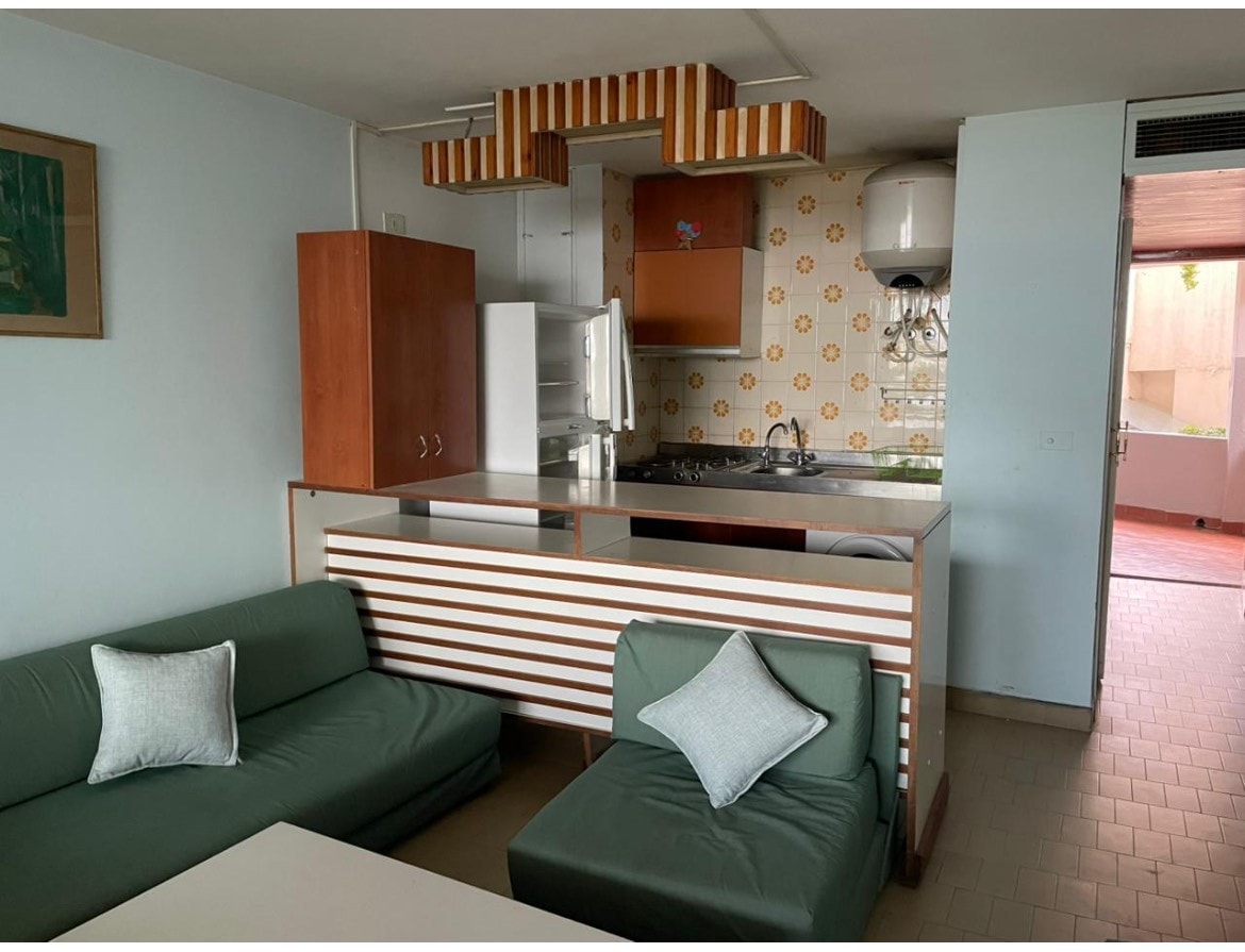 A small living area features a kitchenette with wooden cabinetry and floral tiles. Two green sofas with accent pillows are positioned around a white coffee table, creating a comfortable seating arrangement. A door leads to the outdoor veranda, allowing natural light to brighten the space.