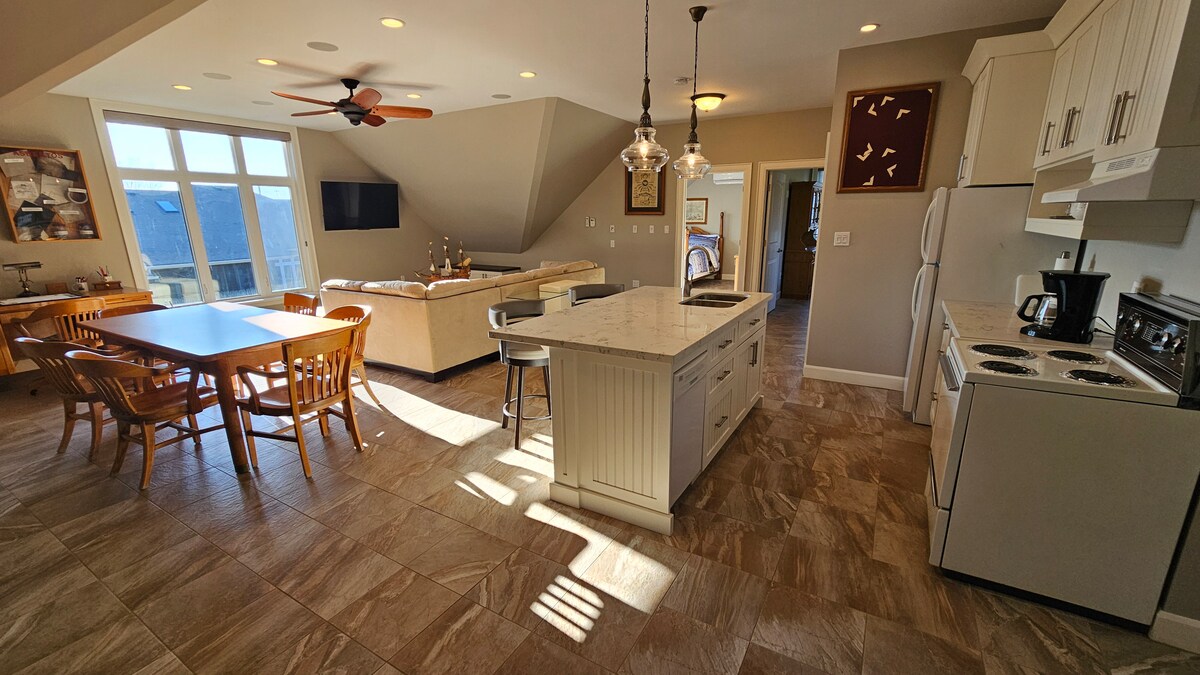 A bright and open kitchen and living area features large windows allowing natural light to fill the space. A central island with seating is surrounded by a dining table and comfortable seating arrangements. Modern appliances and warm tones create an inviting atmosphere.