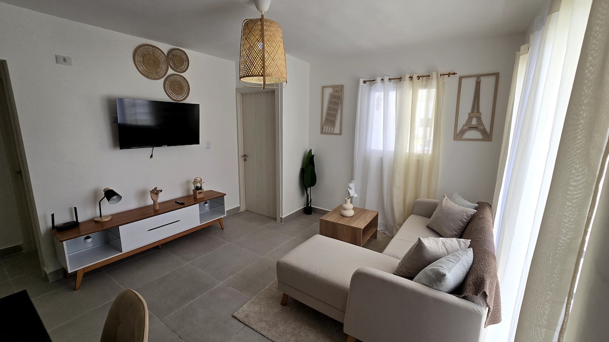 A well-lit living area features a light-colored sofa adorned with soft pillows. A wooden coffee table sits in front, complemented by a modern TV mounted on the wall. Large windows with sheer curtains allow natural light to fill the space, enhancing the minimalistic decor.