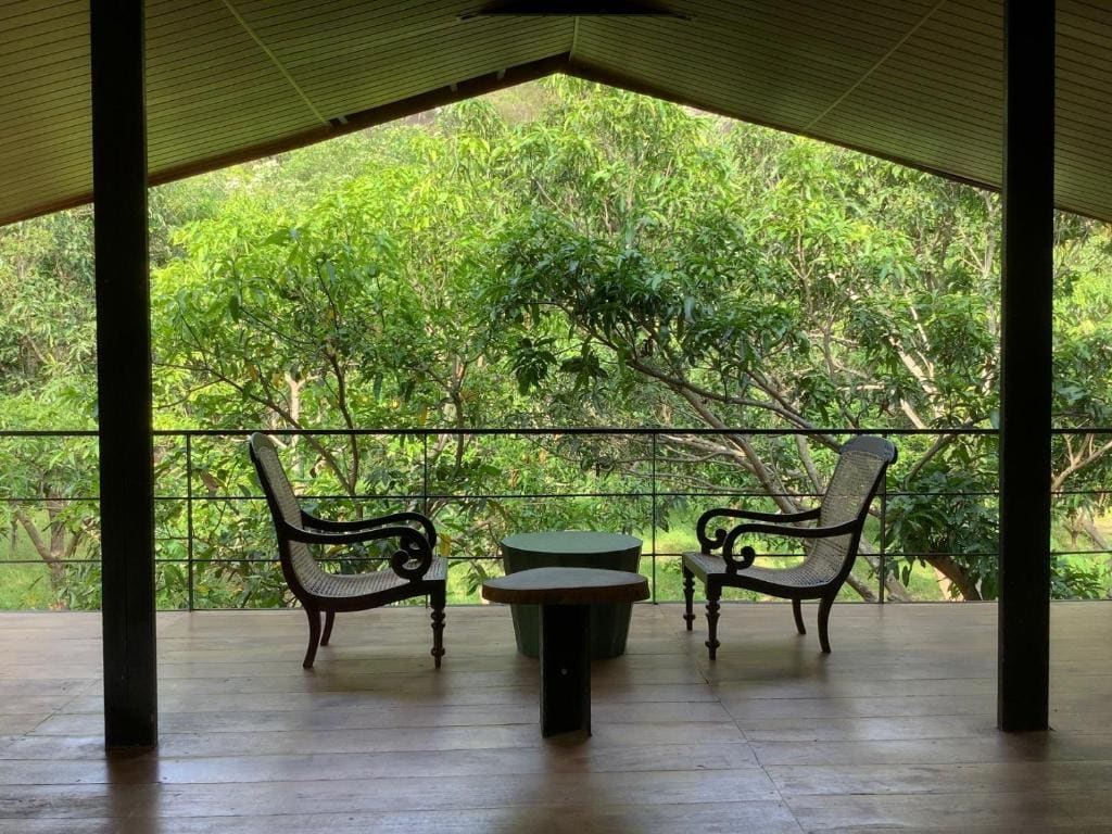 A serene balcony area features two elegant chairs positioned around a round table, overlooking a lush expanse of greenery. The sloped ceiling and wooden flooring contribute to a natural feel, while the railing allows unobstructed views of the surrounding trees.