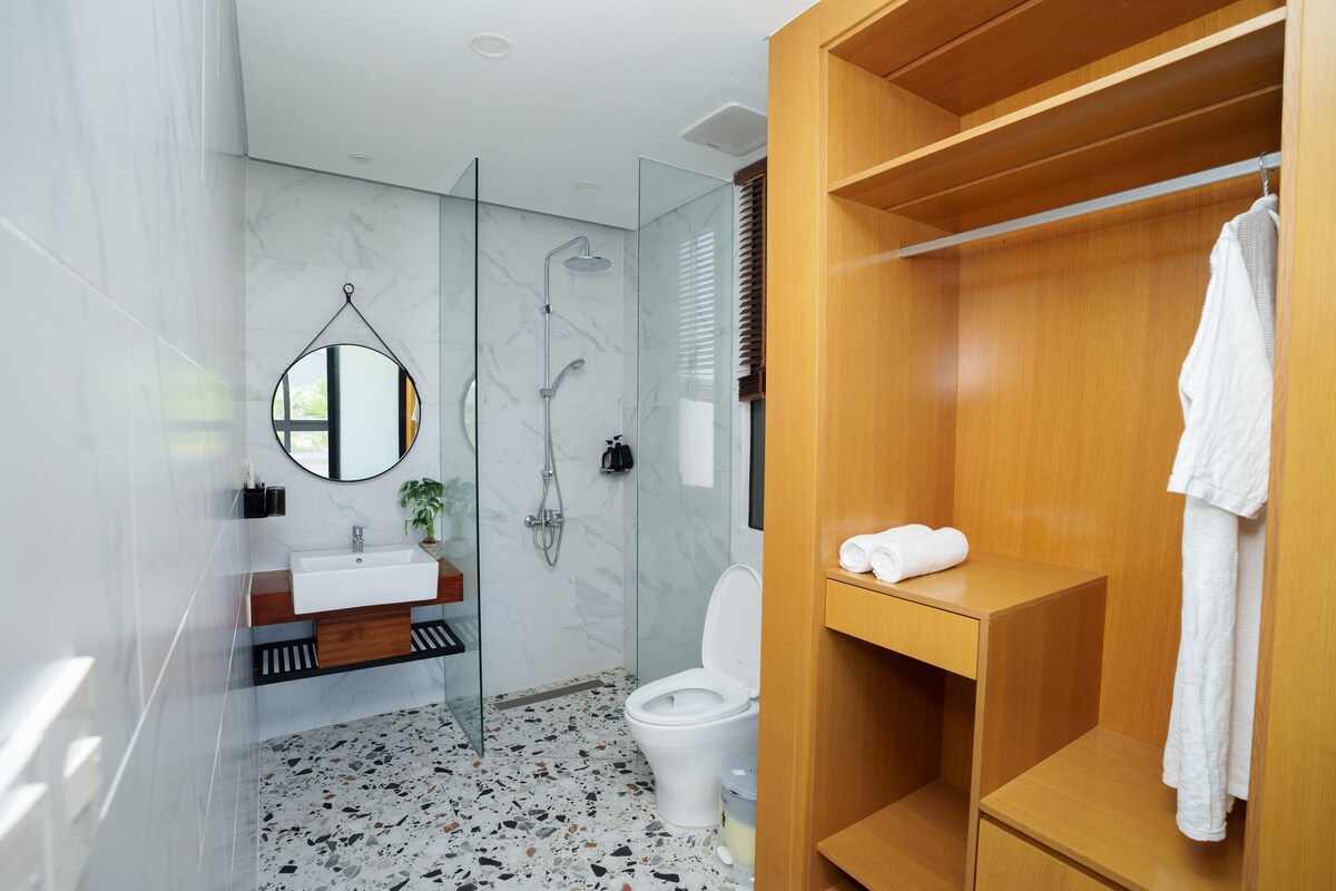 A modern bathroom features sleek marble walls and a spacious shower area with a glass enclosure. A round mirror hangs above a wooden vanity, which includes a small plant. A wooden wardrobe offers a towel and robe, enhancing the functional layout.