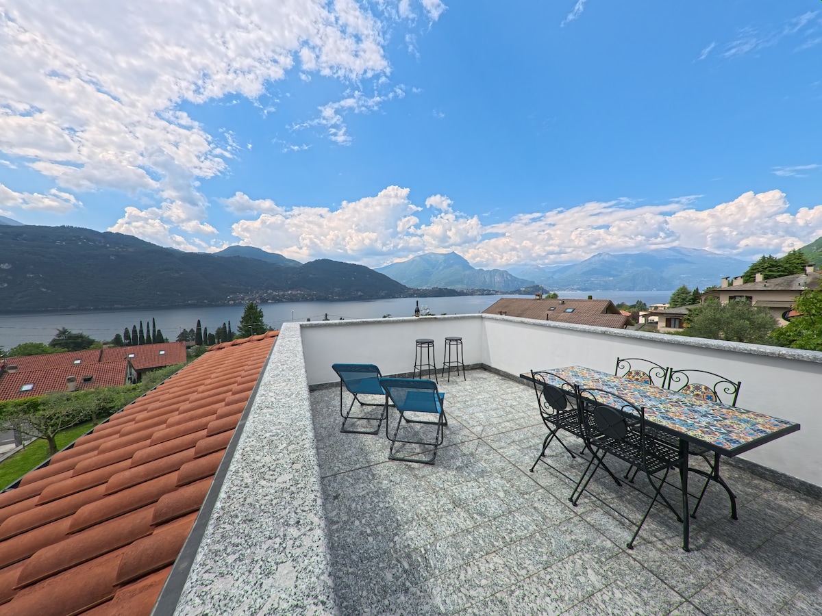 A spacious private terrace is featured, offering views of Lake Como and surrounding mountains. Outdoor furniture includes a colorful dining table with six chairs and two lounge chairs. The terrace is surrounded by a low wall, providing an open feel.