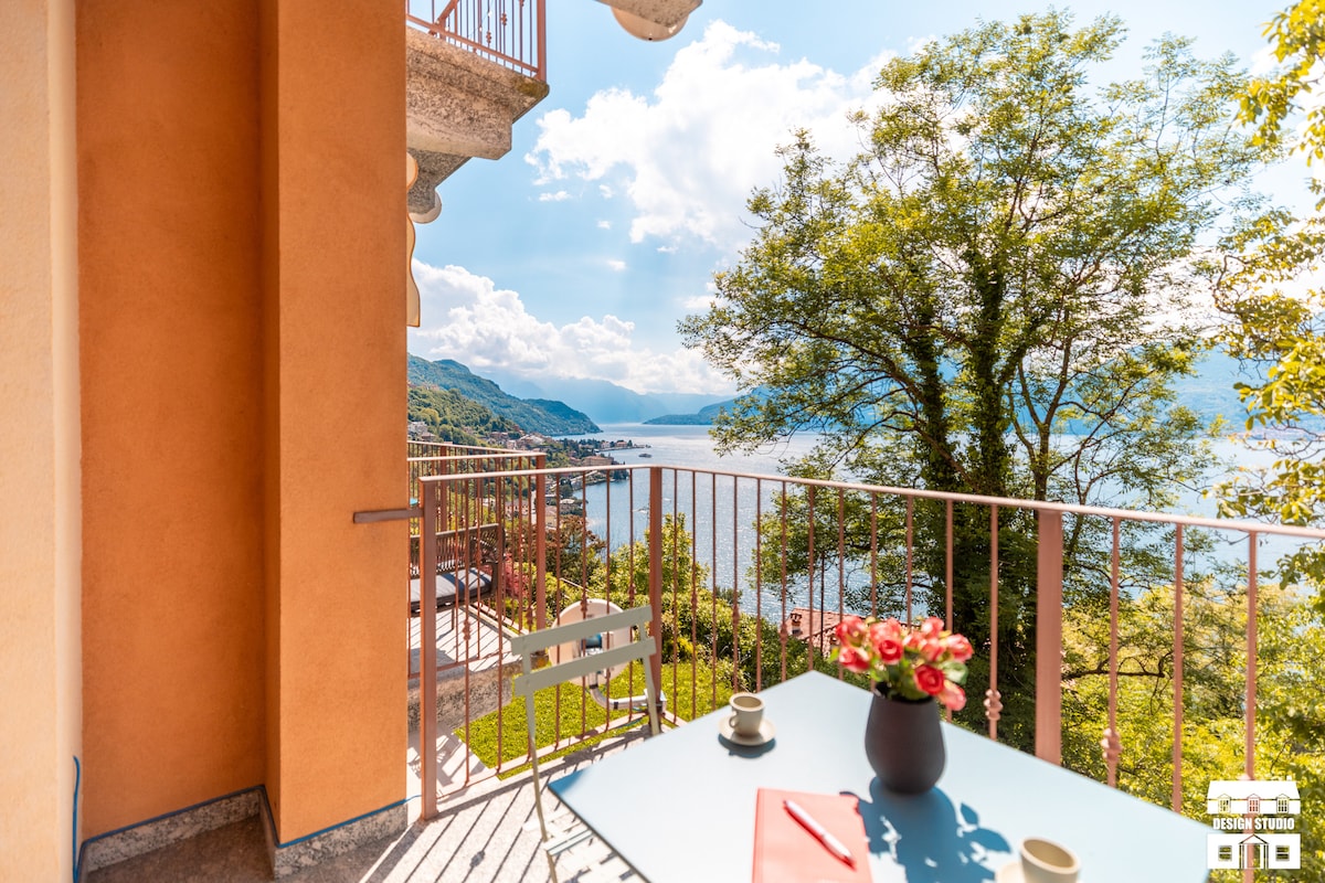 A balcony is visible, featuring a table set with two mugs and a vase of flowers. Verdant trees frame the view of the lake and distant hills. Soft sunlight illuminates the scene, enhancing the serene outdoor space designed for relaxation.