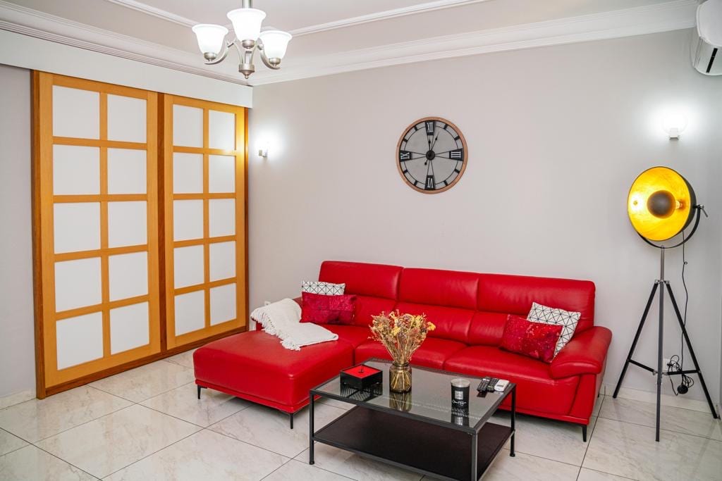 A modern living room is featured with a red sectional sofa and an accompanying coffee table. A wall clock is positioned above the sofa, while a warm floor lamp stands nearby. Light-colored walls and glossy tile flooring create an open and airy feel.