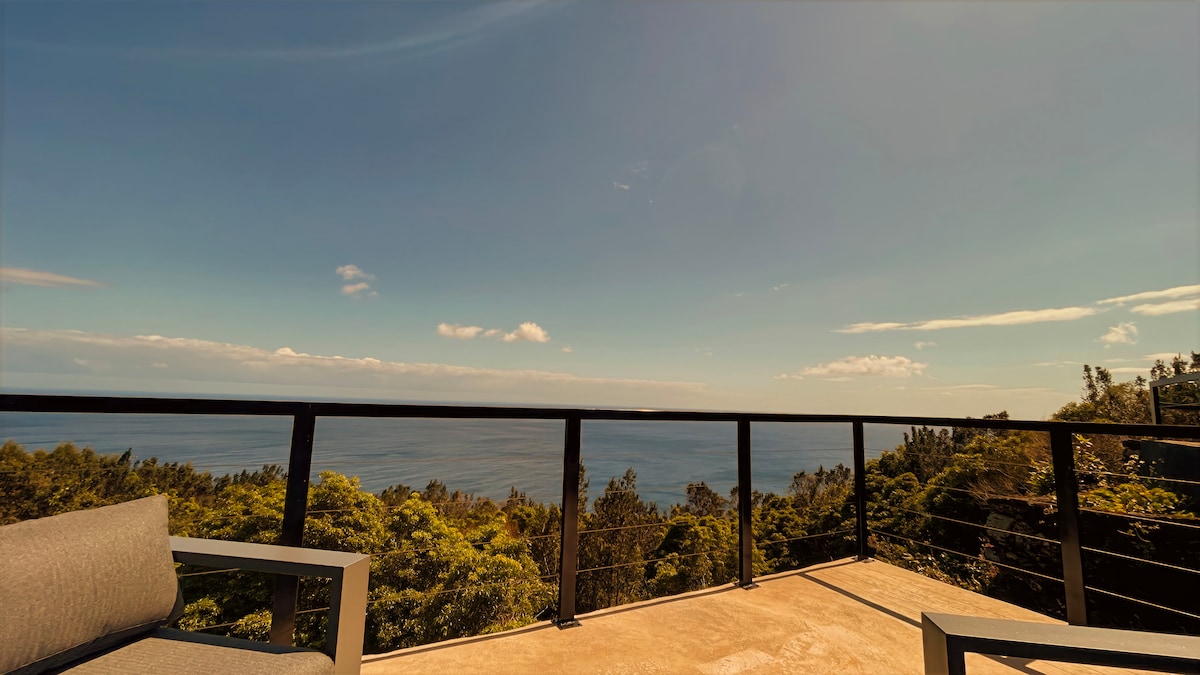 An outdoor terrace is presented with a railing, showcasing expansive ocean views. Two gray lounge chairs are positioned on the concrete surface, surrounded by greenery and open sky. The scene reflects natural light and serenity, inviting moments of relaxation.