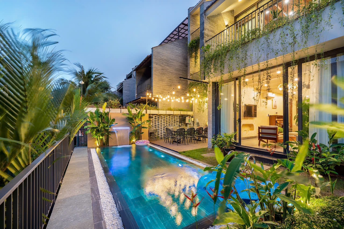 The outdoor area features a private swimming pool surrounded by lush greenery. The villa's contemporary structure showcases a blend of natural elements and modern design. Soft lighting enhances the ambiance, providing a welcoming space for relaxation or social gatherings.