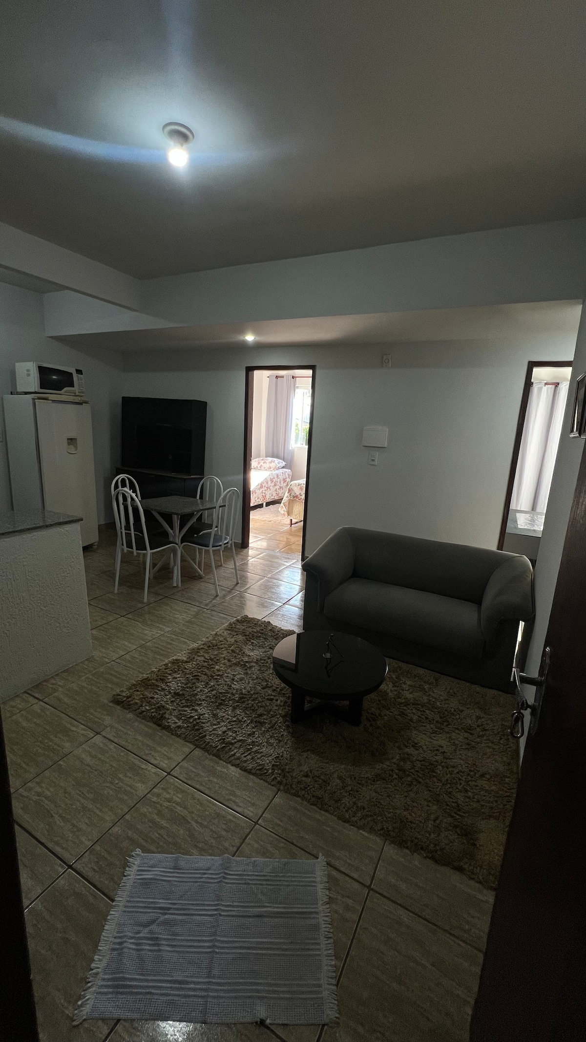 A living space is showcased, featuring a gray sofa and a small round table with chairs. A light rug is positioned on the tiled floor. Access to a bedroom area is visible through an open door, while the kitchenette and a television are noted in the background.
