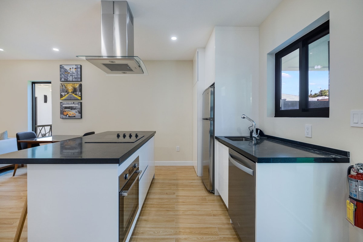 A modern kitchen features an island with a smooth, dark countertop and a built-in stovetop. Stainless steel appliances, including a refrigerator and a sleek sink, are visible. Light wood flooring complements the bright space, while large windows allow natural light to fill the room.