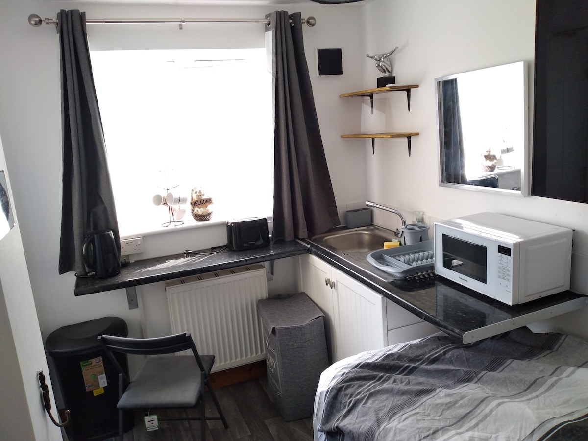 A kitchenette studio is showcased, featuring a workspace with a black countertop and a sink. A microwave and kettle are positioned nearby. The window is adorned with grey curtains, and a chair is placed next to the desk. A cozy sleeping area is visible in the corner.