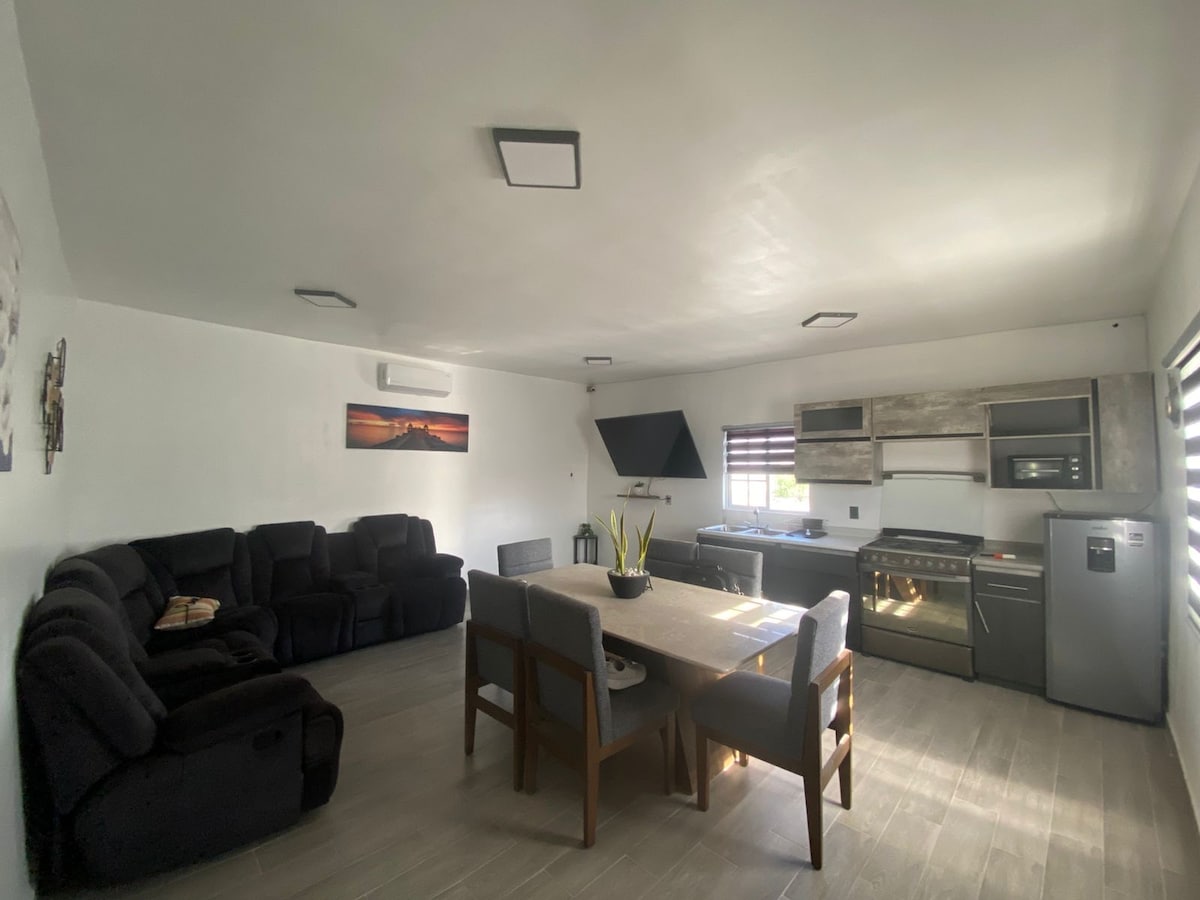 An open living and dining area showcases a dark sectional sofa and a wooden dining table surrounded by chairs. A sleek kitchen with modern appliances, including a stove and refrigerator, is visible in the background. Soft overhead lighting brightens the space.