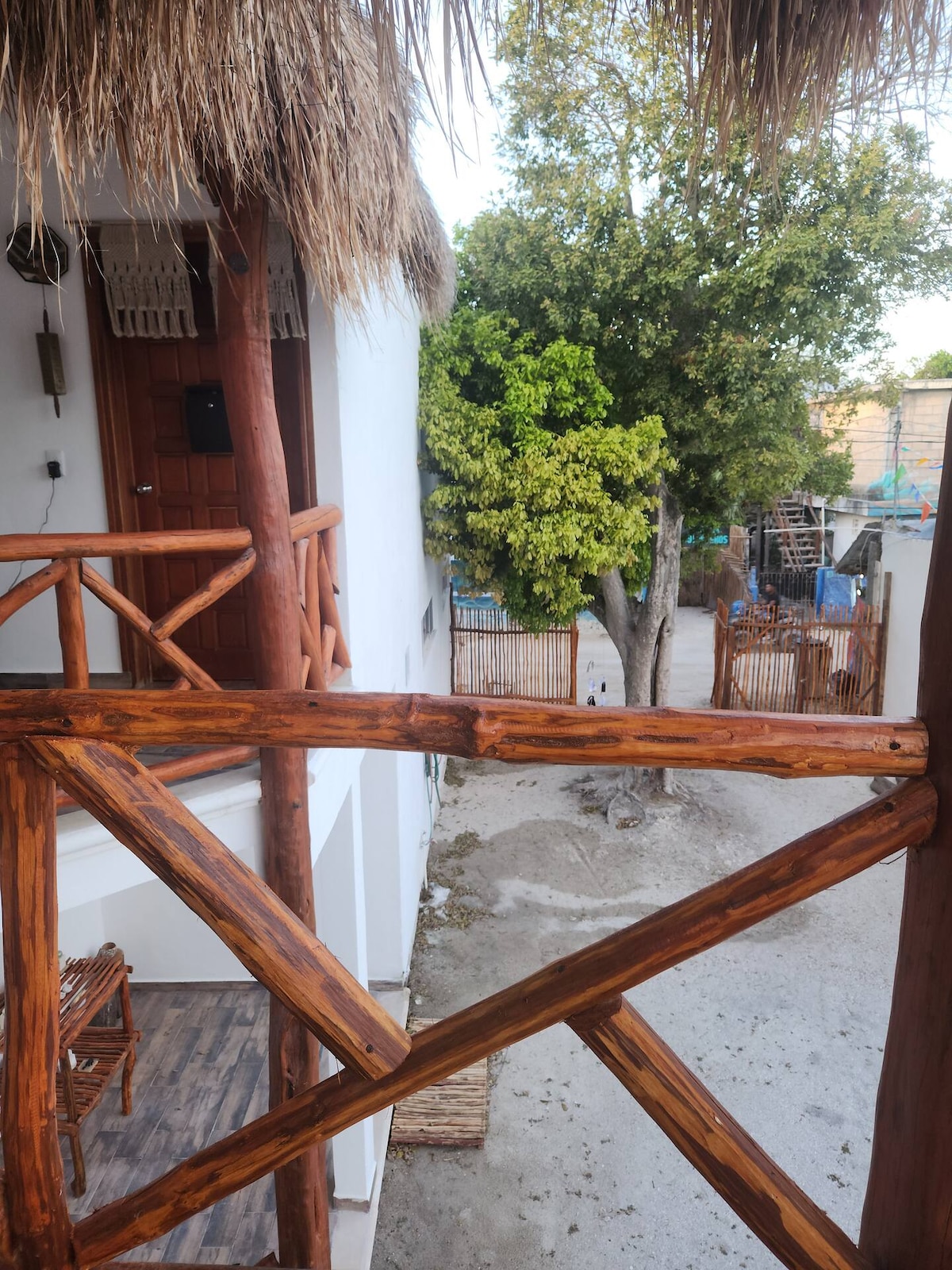 A wooden balcony railing frames a view of the pathway below, lined with greenery. A tree stands prominently on the left, while various architectural elements are visible in the background, showcasing a blend of natural and constructed aspects of the vicinity.
