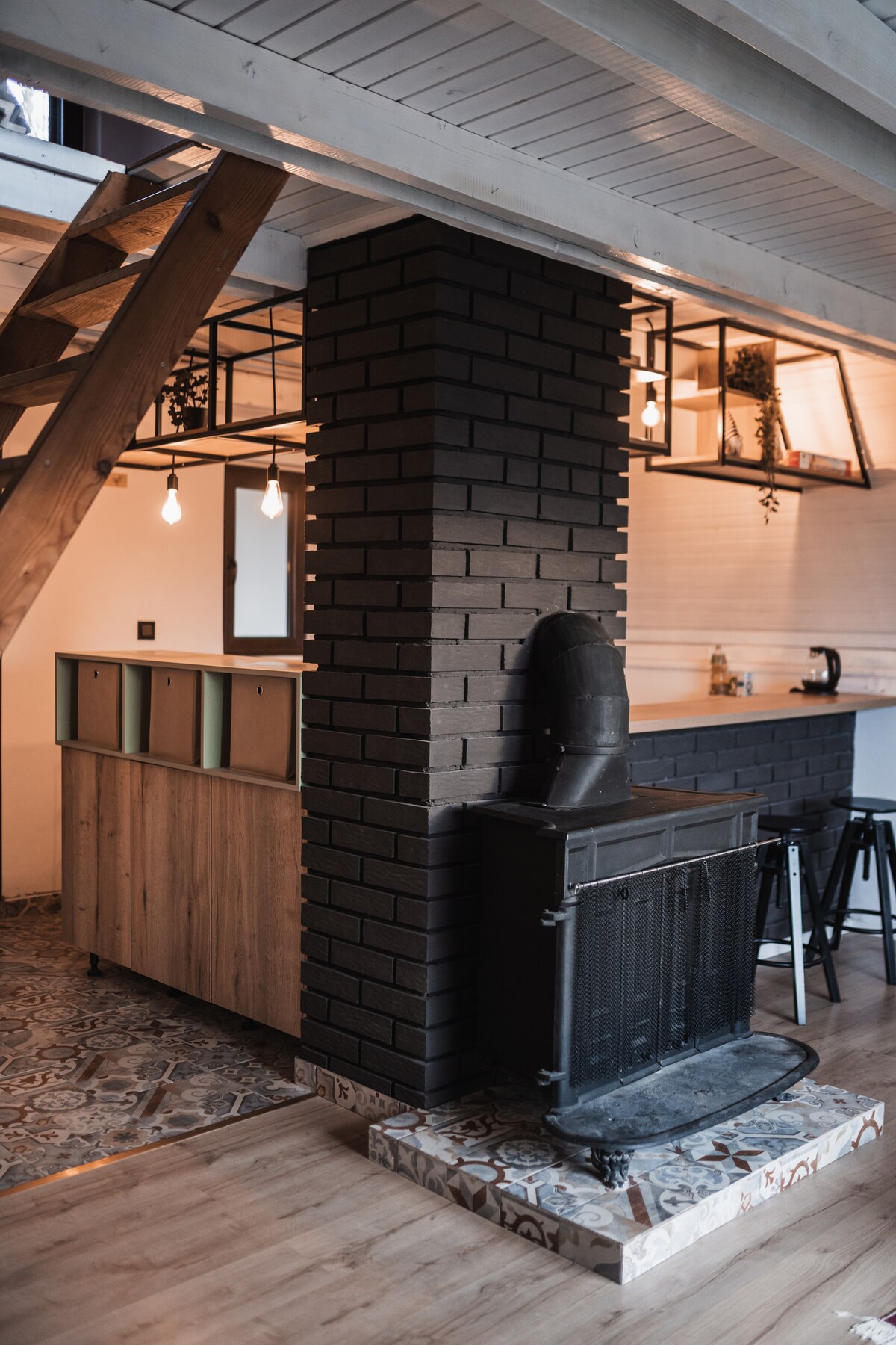 An open-plan living area features a central brick column with a vintage stove, surrounded by a kitchen island with a mix of wooden and shelving units. Pendant lights hang above, and a staircase with wooden steps leads to the upper level, emphasizing the space's inviting design.