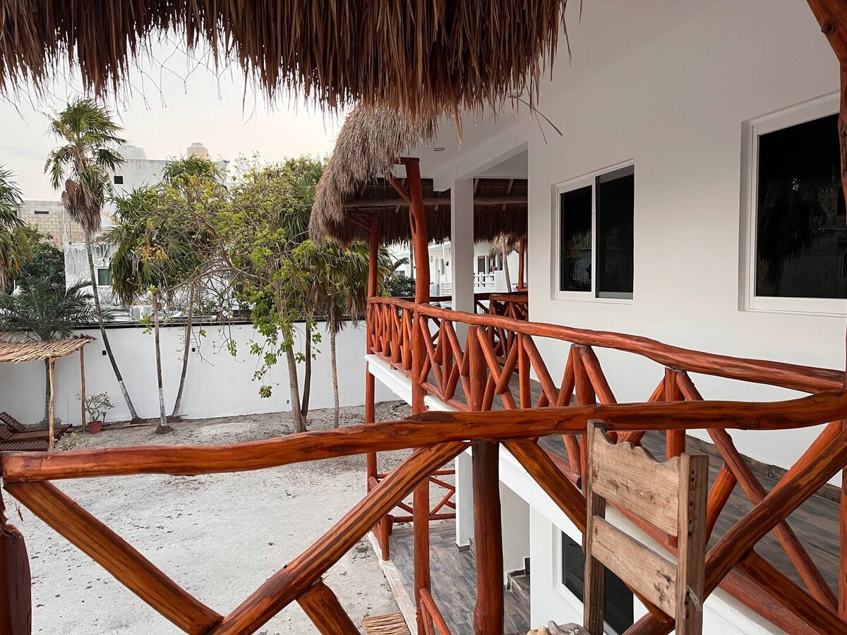A wooden balcony with a straw roof extends from the building, showcasing natural wood railings and the surrounding garden area. Large windows with neutral trim are visible, and a serene outdoor space is presented, inviting moments of relaxation.