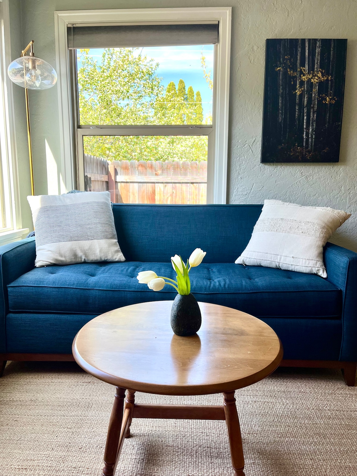A cozy seating area features a blue sofa adorned with two decorative pillows. A small round table sits in front, hosting a dark vase filled with white tulips. Natural light streams through a nearby window, revealing greenery outside.
