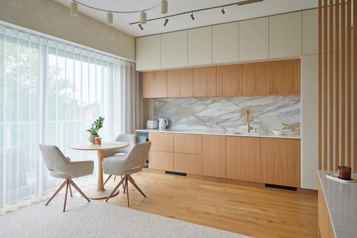 A modern kitchen area features natural wood cabinetry and a marble backsplash. A round dining table with two light grey chairs is positioned near the large window, which is dressed in sheer white curtains, allowing ample natural light to fill the space.