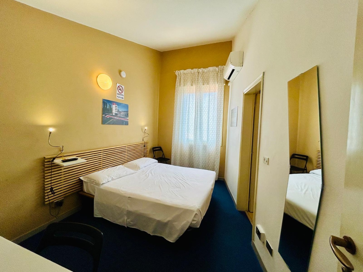 A well-lit sleeping area features a spacious bed with crisp white linens, complemented by a wooden headboard. Natural light enters through sheer curtains, highlighting the warm beige walls. A full-length mirror adds depth to the space, while a simple desk and chair are positioned nearby.