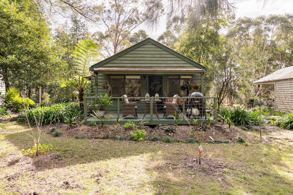 A quaint cabin is surrounded by lush greenery and native plants. The front porch features seating, inviting relaxation while overlooking the tranquil outdoor space. Large windows allow natural light to fill the interior, blending the cabin harmoniously with the scenic landscape.