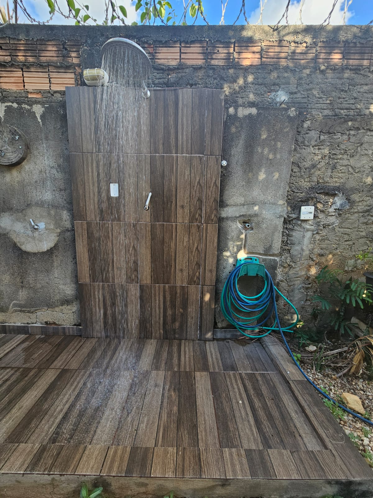 An outdoor shower is featured, designed with wooden panels and a stone backdrop. Water flows from a showerhead above, creating a refreshing experience. A garden hose is seen coiled nearby, and natural greenery adds a tranquil setting.