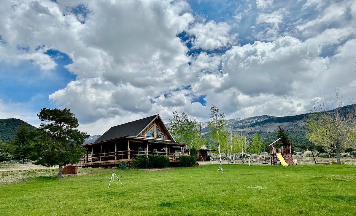 A charming cabin sits on a spacious green lawn, framed by trees and surrounded by rolling hills. The structure features a large front porch and a visible playset, while dramatic clouds drift across the sky, adding character to the serene landscape.