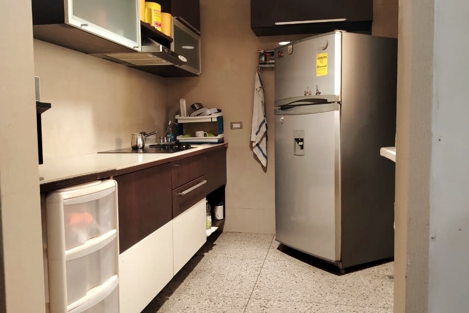 The kitchen features modern cabinetry and a silver refrigerator on the right. Countertops are clean and spacious, with a sink and various utensils visible. Clear storage containers and a small shelf can be seen, providing organization and functionality.