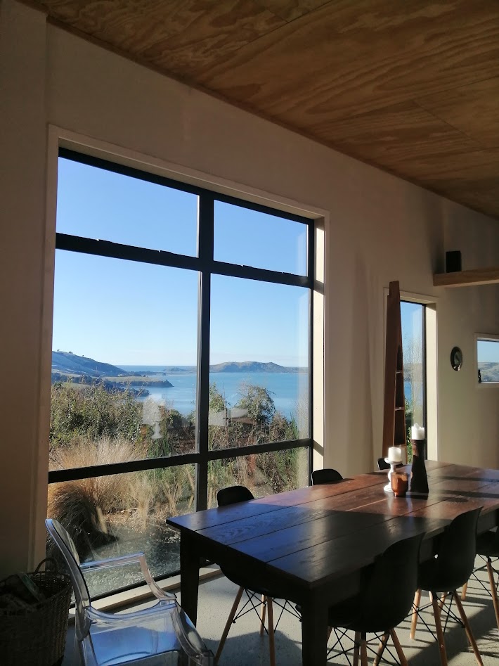 A dining area is presented with a large wooden table surrounded by several chairs. A large window offers unobstructed views of the water and surrounding landscape, allowing natural light to flood the space. The interior features a clean, modern aesthetic.