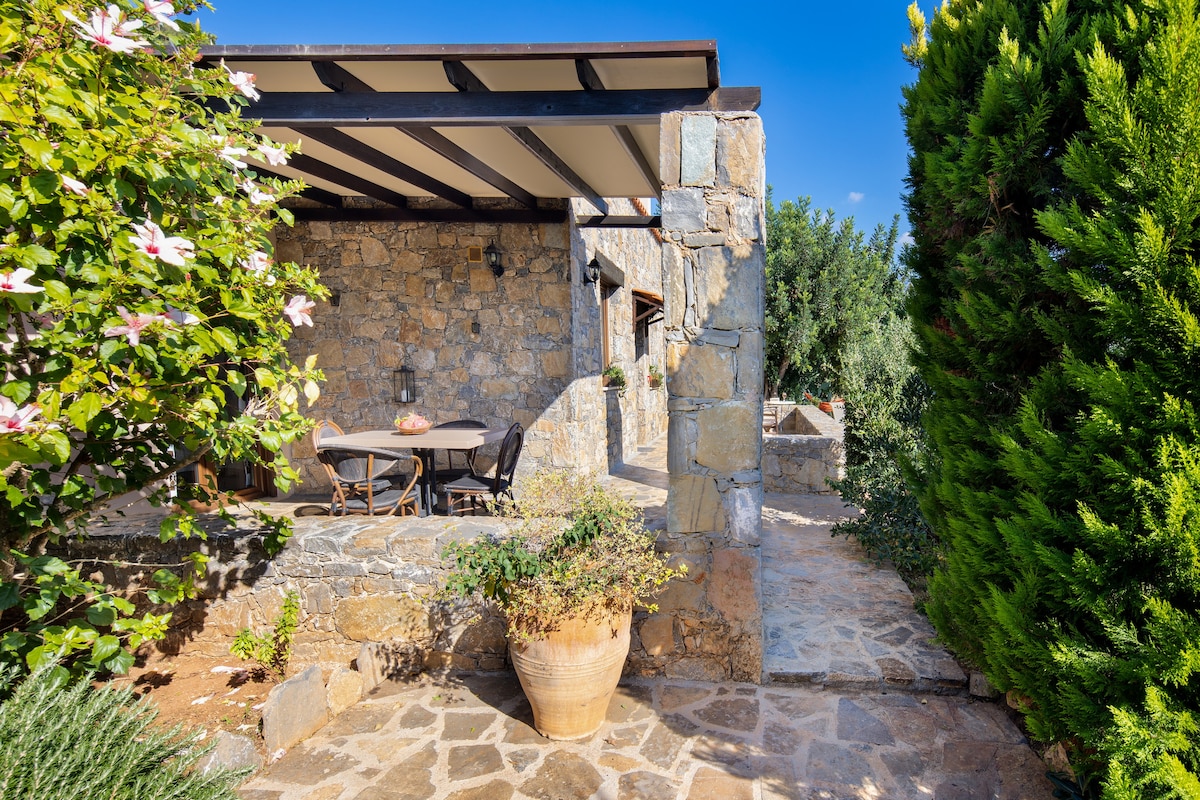 The image depicts a stone exterior of a villa, featuring a covered terrace with a dining table. Lush greenery surrounds the area, with flowering plants and tall hedges providing privacy. A stone pathway leads towards the entrance, creating a welcoming atmosphere.