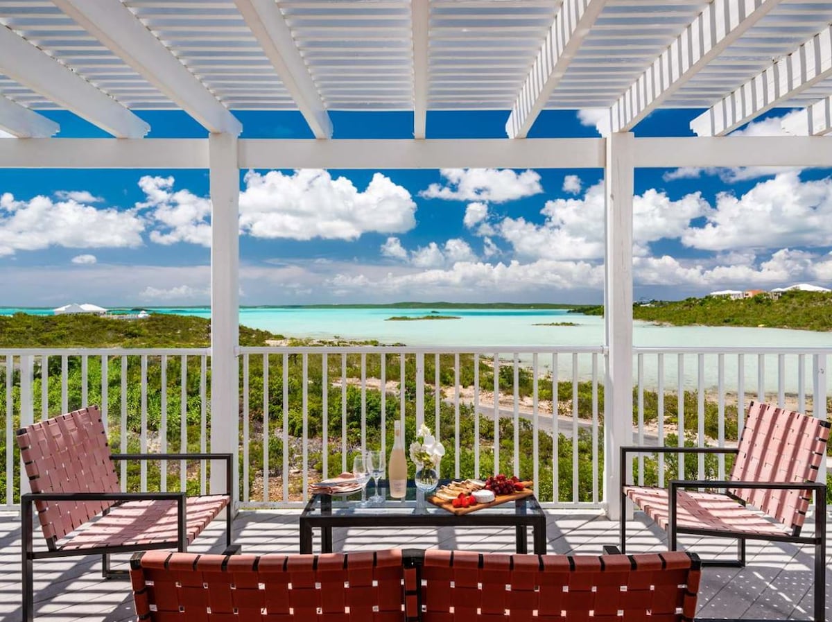 An outdoor balcony features a seating arrangement with four woven chairs and a small table. A scenic view of the water and lush landscape is visible, framed by the white wooden railing and overhead pergola. Soft clouds float in a bright blue sky.