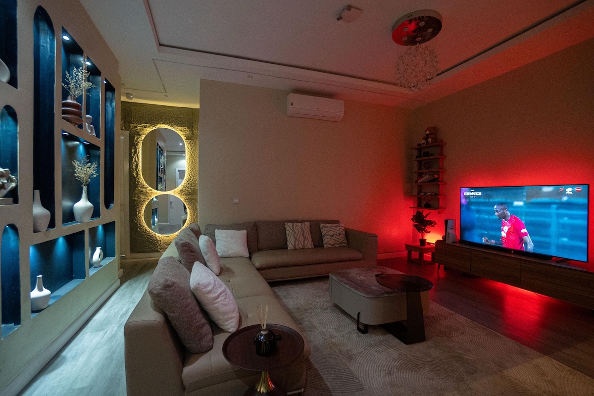 A spacious living area features a large sectional sofa, accented with decorative pillows. A modern coffee table sits in front. A wall-mounted television displays a sports game, while ambient lighting casts a warm glow over the room, highlighting a decorative shelving unit.