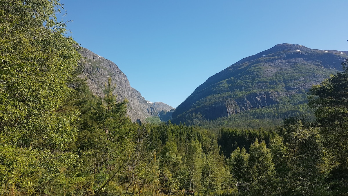 A serene landscape displays two majestic mountains framing a lush valley. Various trees and greenery extend across the foreground, while the clear blue sky and bright sunlight create a peaceful ambiance.
