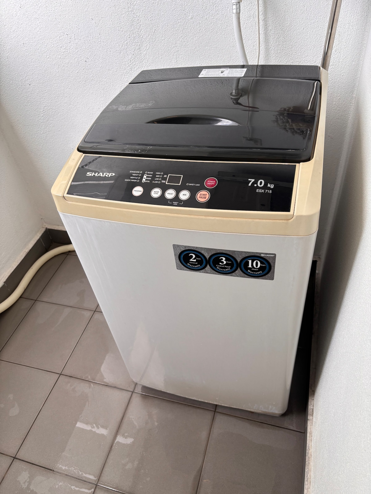 A top-loading washing machine is positioned in a corner. It features a sleek design with control buttons visible on the front panel. The clean, tiled floor reflects light, and the surrounding walls are simple and unobtrusive.
