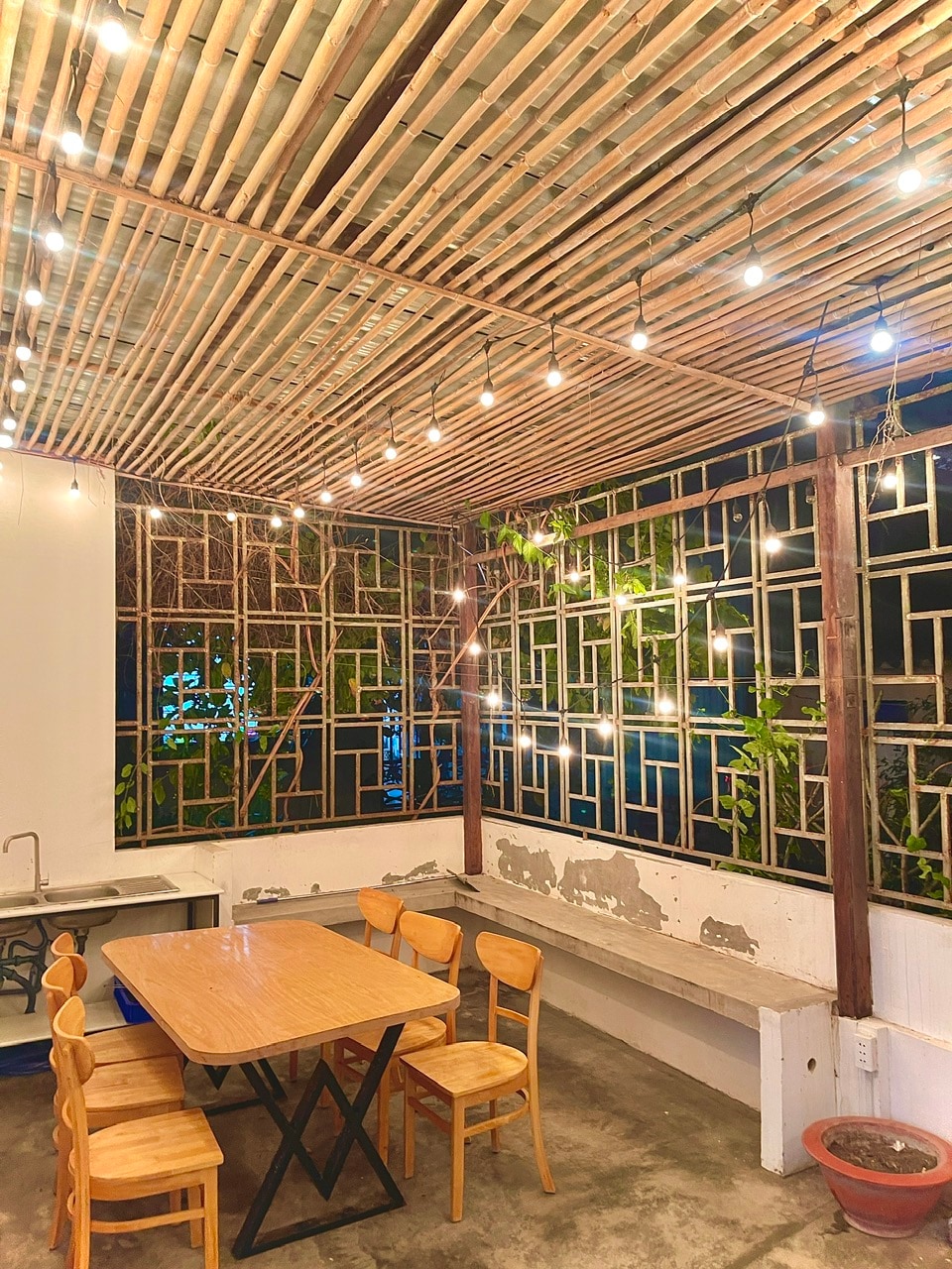 An open dining area features a wooden table surrounded by six chairs. The ceiling is adorned with hanging lights, while large windows allow views of greenery outside. A sink is located in the kitchen area, creating a sense of connection with the outdoor environment.