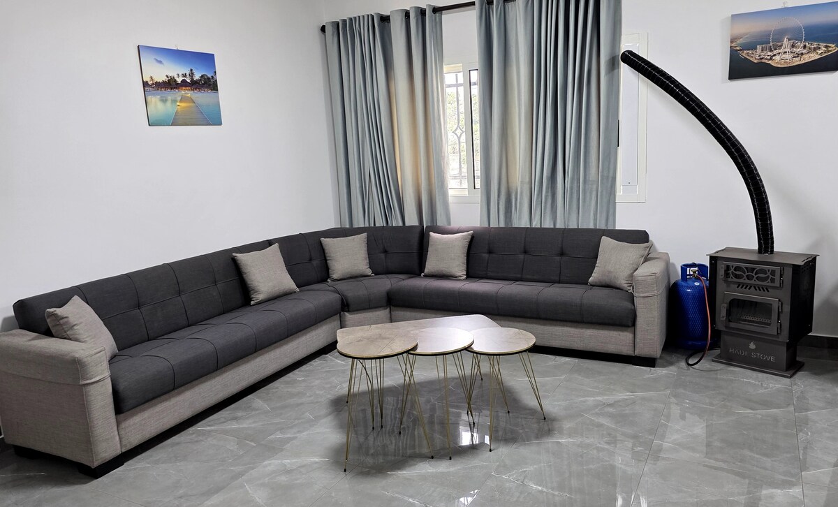 A spacious living area features a modern sectional sofa in neutral tones, complemented by decorative cushions. Two small, round tables sit on polished gray tile flooring. Natural light filters through floor-length curtains, and a heating unit is visible in the corner near a gas cylinder.