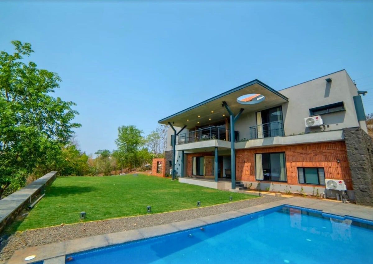 A modern villa with a sleek design is set against a clear blue sky. The structure features large windows and a spacious terrace. A private pool reflects the surroundings, while manicured green grass complements the earthy brick facade.
