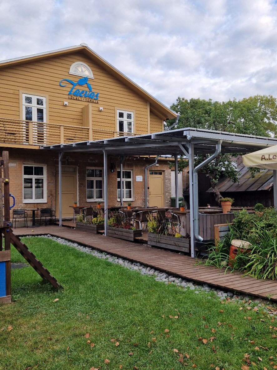 An exterior view of the guesthouse features a two-story wooden structure with a light yellow façade and blue signage. A wooden deck extends from the entrance, surrounded by greenery and decorative plants. Outdoor seating is provided under a covered area, inviting relaxation.