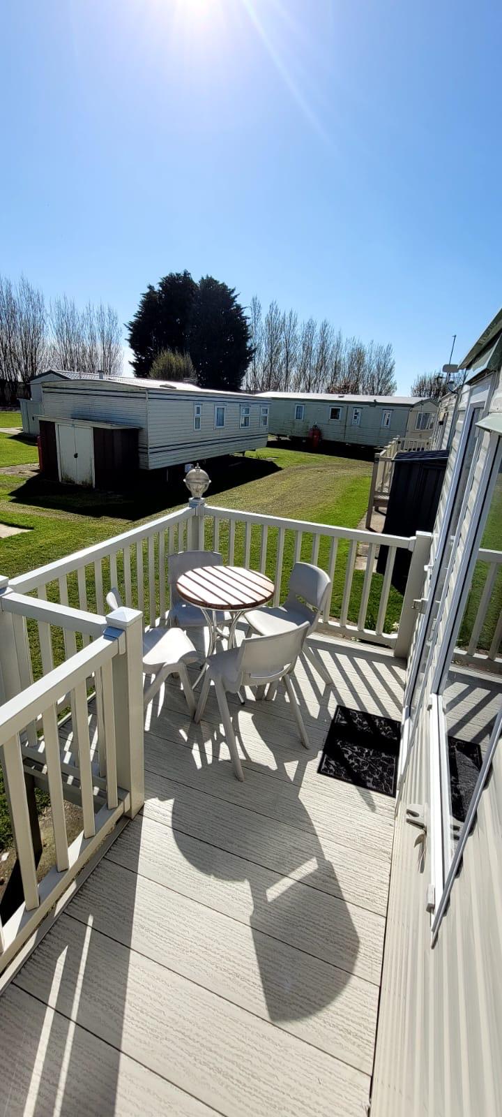 A private decking area presents a round table accompanied by two chairs, surrounded by well-maintained green grass. Nearby caravans are visible, set against a bright blue sky, suggesting a sunny and open atmosphere.