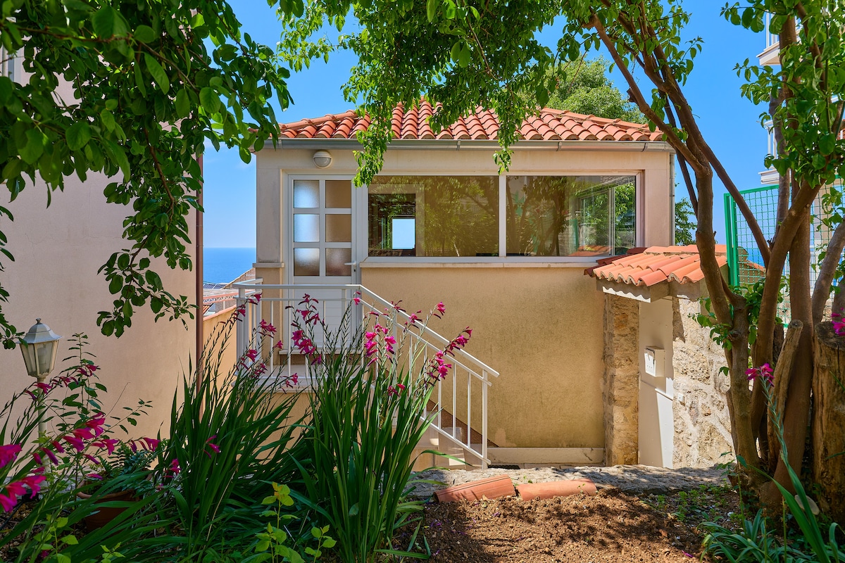 The exterior of the apartment is showcased, featuring a light-toned structure with a red-tiled roof. Lush greenery surrounds the pathway leading to the entrance, while vibrant pink flowers add color. The ocean is visible in the background, framed by branches and leaves.
