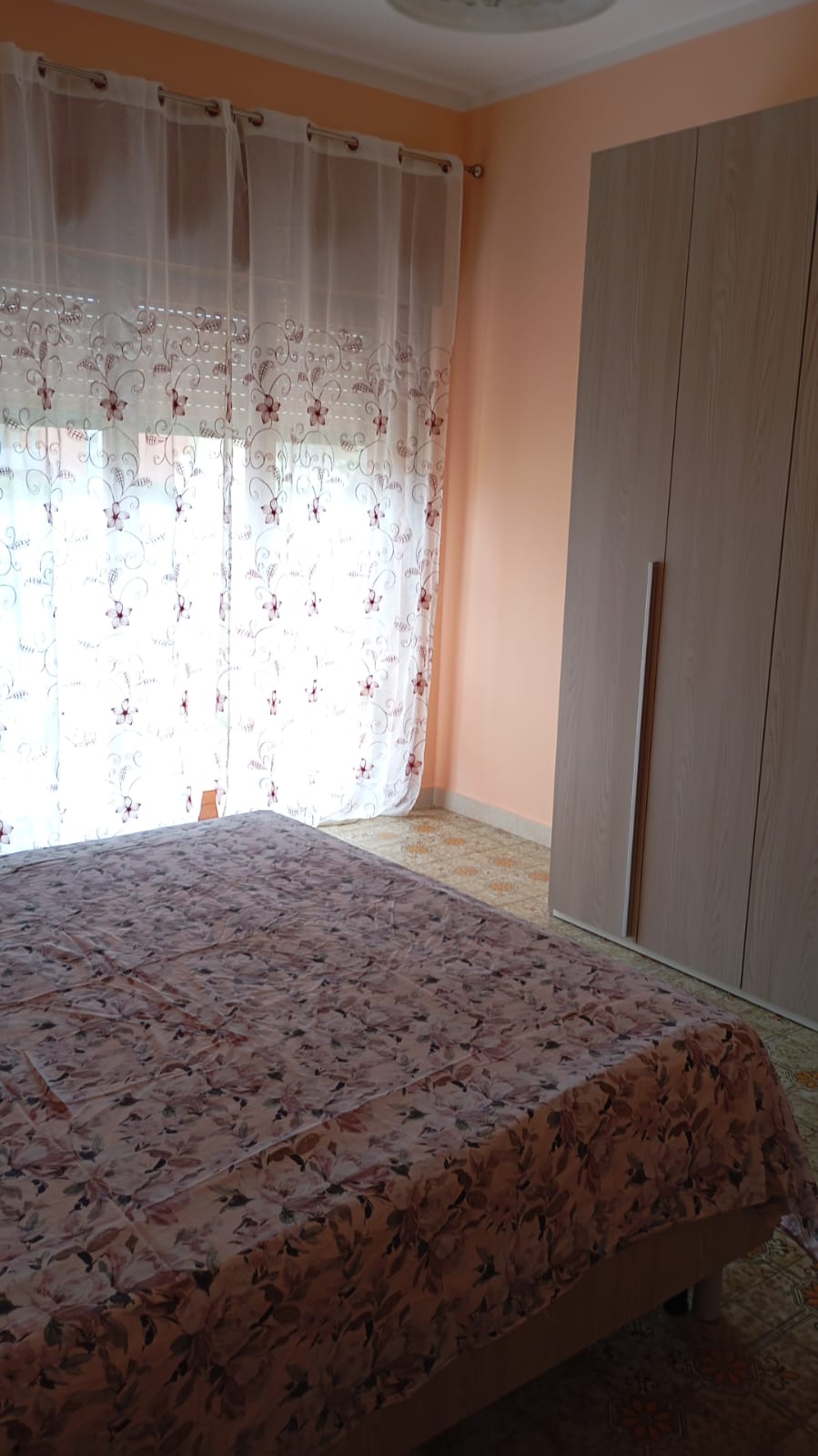 A cozy bedroom is featured with a floral-patterned bedspread covering a spacious bed. Light filters through sheer curtains adorned with floral designs, illuminating the room. A large wardrobe stands against one wall, complementing the soft peach-colored walls and patterned floor.