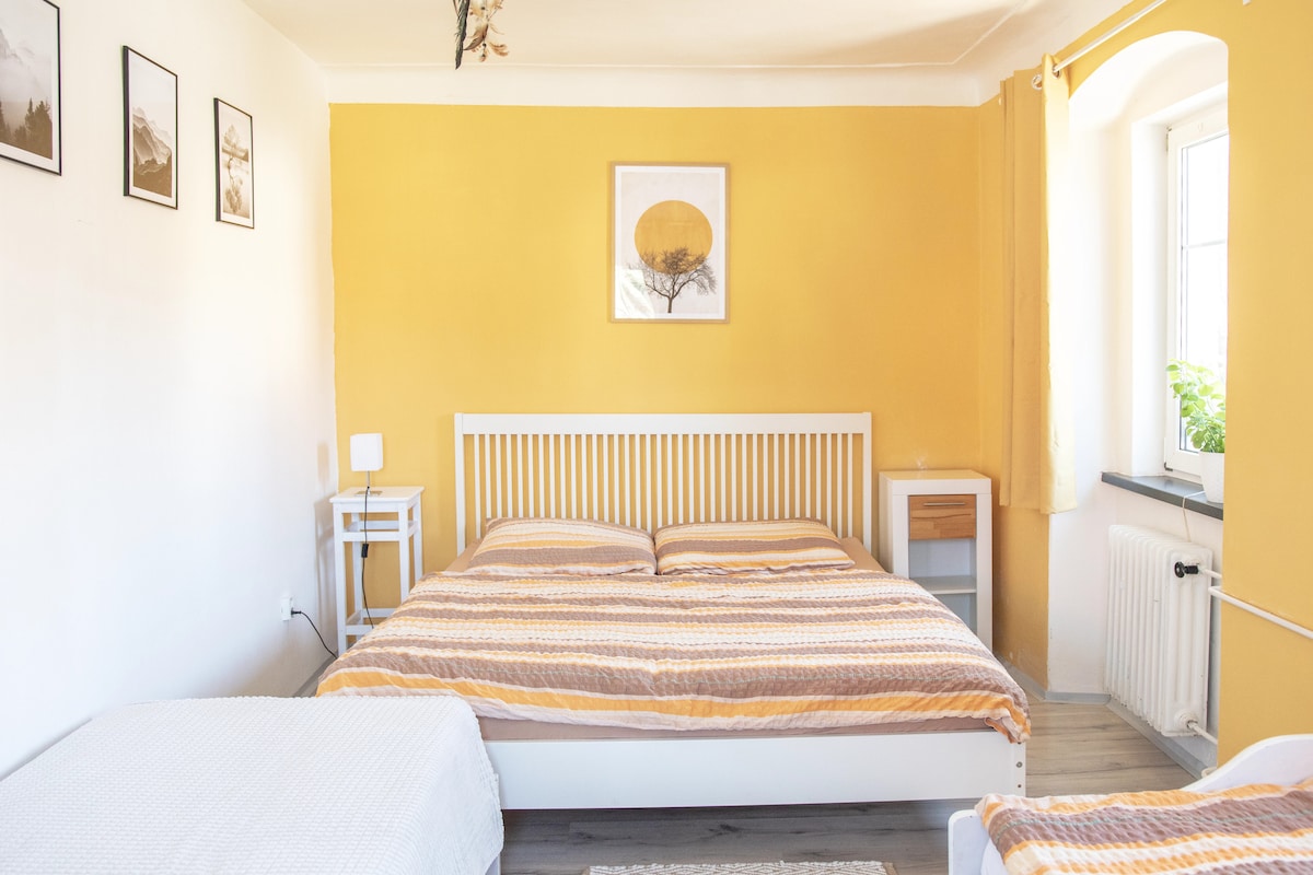 A bright sleeping area features a spacious bed with striped bedding, complemented by a smaller bed positioned nearby. The walls are painted a warm yellow, and framed photographs adorn the wall. Natural light flows through the window, enhancing the inviting atmosphere.