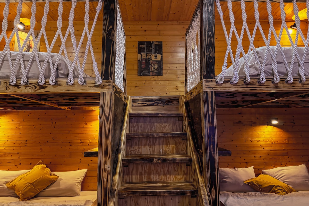 A cozy multi-level sleeping area is framed with wood paneling, featuring two lofted beds connected by a rustic rope railing. A staircase leads to the upper level, while soft lighting creates a warm ambiance. Plush yellow pillows are positioned on the beds, inviting relaxation.