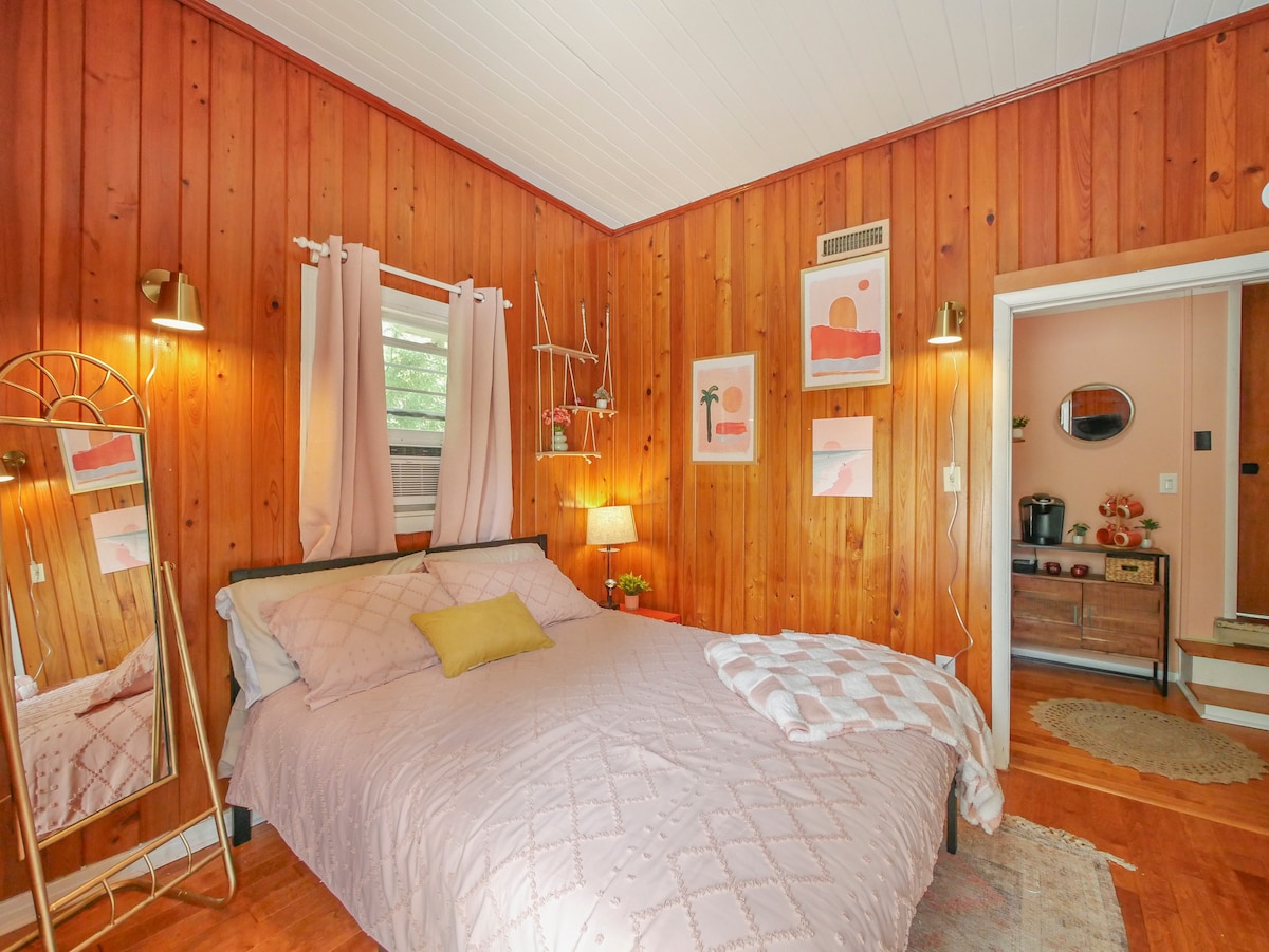 A cozy bedroom features warm wooden paneling and a queen-sized bed with light linens. A small accent pillow adds a touch of color. A full-length mirror stands beside the bed, and soft lighting is provided by a bedside lamp. Decorated wall art complements the inviting space.
